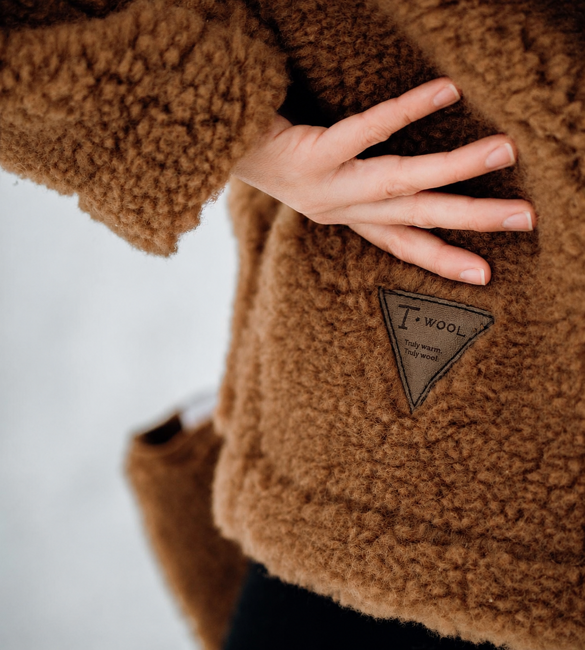 En kvinnelig hånd som viser frem en logo på en brun ulljakke der det står skrevet T-wool.